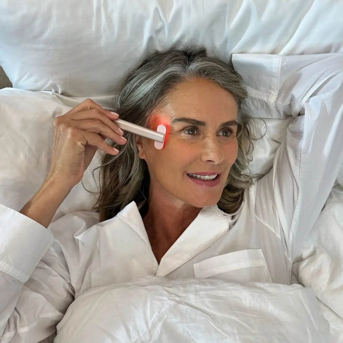 An image of an elderly woman laying in bed using her Solawave red light therapy wand on her face