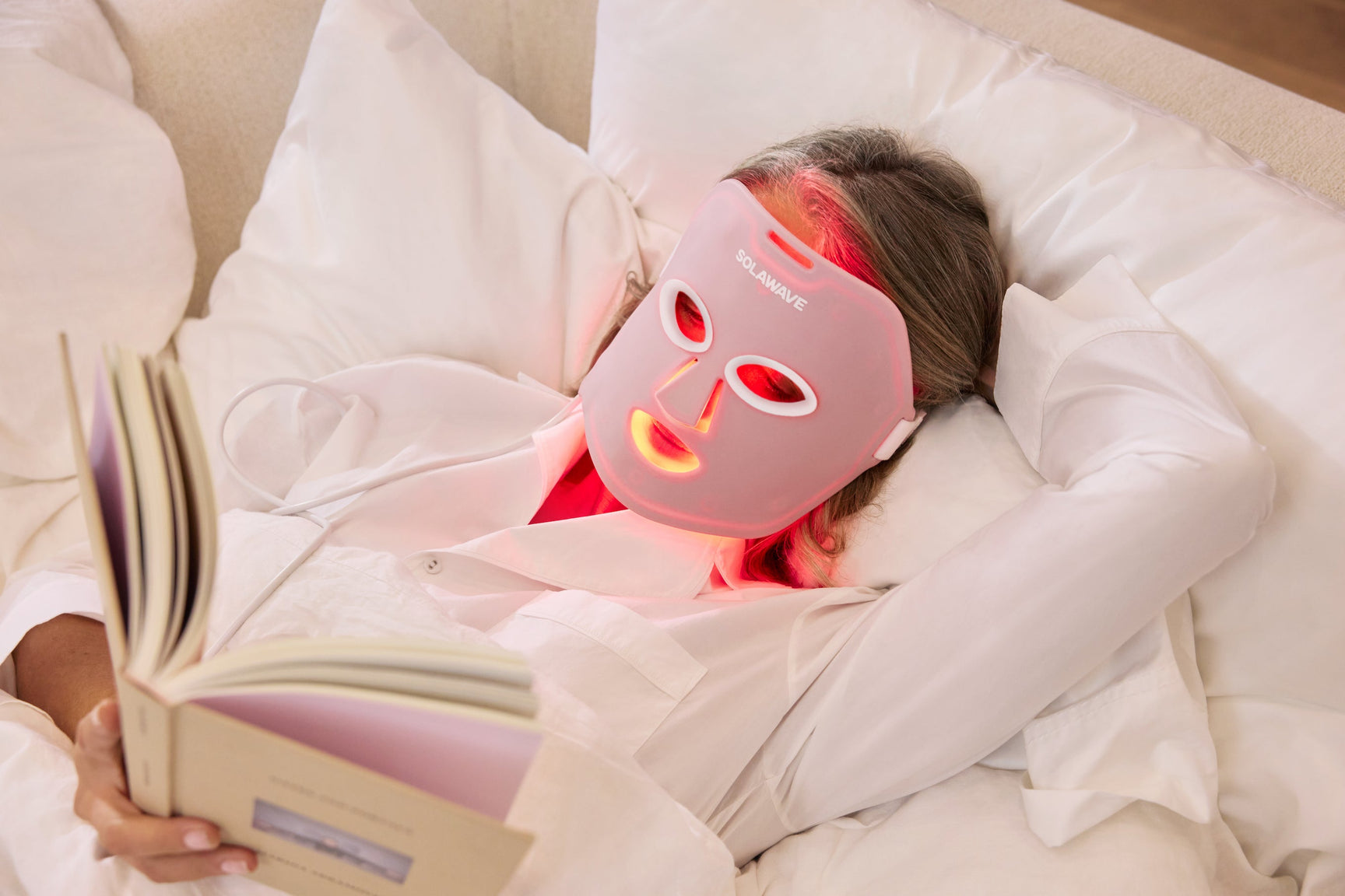 An image of a woman using a red light therapy mask at night reading a book in her bed