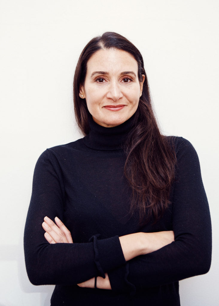 A woman with long brown hair stands against a plain white background. She is wearing a black turtleneck sweater and has her arms crossed, smiling softly at the camera.