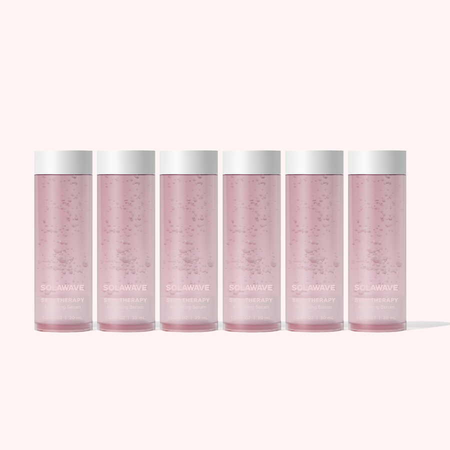 Six translucent pink bottles with white caps, part of the Solawave Skin Therapy Activating Serum Refill 6-Pack, are lined up against a pale background. The bottles read Solawave and THERAPY, suggesting anti-aging benefits. The serum inside reveals tiny bubbles.