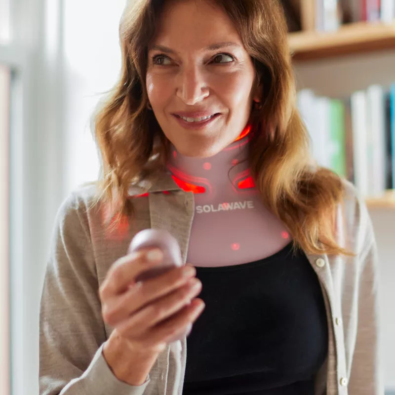 Woman wearing the Solawave Neck & Chest Pro Light Therapy Mask with red light therapy, holding its remote