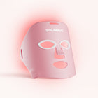The Solawave Wrinkle Retreat Light Therapy Face Mask is pink with cutouts for the eyes, nose, and mouth. It emits a subtle red glow using red and near-infrared light therapy against a soft off-white background for anti-aging treatment.