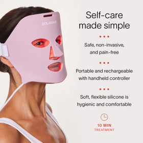A person uses a pink LED mask from Solawaves Wrinkle Retreat Face Mask & LightBoost Cream Kit to boost collagen. It emits red light and features text: Self-care made simple. Safe, non-invasive, pain-free. Portable, rechargeable, soft silicone. 10 min treatment.